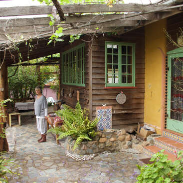 Une femme se tient sur un patio rustique en pierre entouré de plantes, à côté d'une maison en bois dont les fenêtres sont bordées de vert, sous une pergola en bois avec des plantes grimpantes.