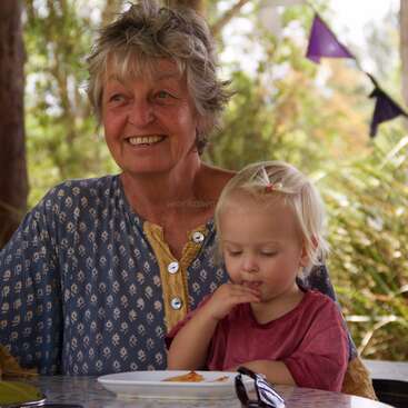 An elderly woman with gray hair smiles warmly, sitting next to a blonde toddler eating at a table outdoors. Colorful bunting hangs in the background. Cozy scene.