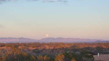 A lua cheia surge acima das montanhas distantes, lançando um brilho suave sobre uma paisagem desértica com arbustos secos e vegetação esparsa, sob um céu em tons pastéis.