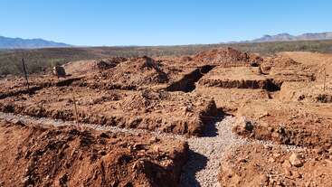 A imagem mostra o canteiro de obras de uma fundação com valas recém-cavadas em solo seco e avermelhado, cercado por vegetação rasteira e montanhas ao longe, sob um céu azul claro.