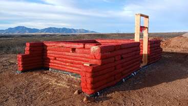 Esta imagem mostra as fases iniciais da construção de uma casa de sacos de terra, com sacos vermelhos empilhados para formar as paredes, uma moldura de porta de madeira e um cenário desértico montanhoso ao fundo.