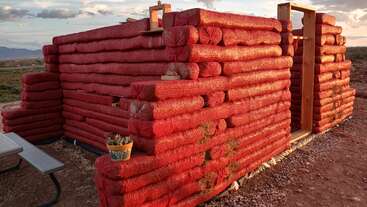 Um prédio em construção feito de sacos de malha vermelhos cheios de terra. Tem molduras de madeira para as portas, um banco e vasos de plantas na parede.