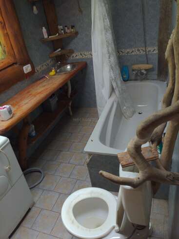 This rustic bathroom features a wooden counter, sink, bathtub with curtain, toilet, washing machine, shelves with toiletries, and unique natural wood accents throughout the space.