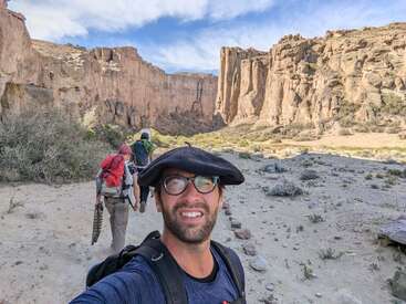 Un homme portant des lunettes et un chapeau prend un selfie lors d'une randonnée avec des amis dans un magnifique paysage de canyon rocheux sous un ciel partiellement nuageux.