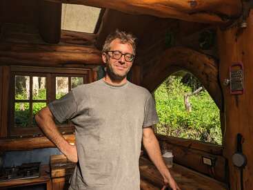 Un homme à lunettes se tient à l'intérieur d'une cabane rustique en bois, la lumière du soleil entrant par les fenêtres, entouré de verdure à l'extérieur. Il semble détendu et satisfait dans un cadre naturel.
