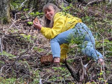 Une femme vêtue d'une veste jaune est en train de butiner sur une colline boisée, tenant une morille et souriant, avec un panier posé sur le sol à proximité.