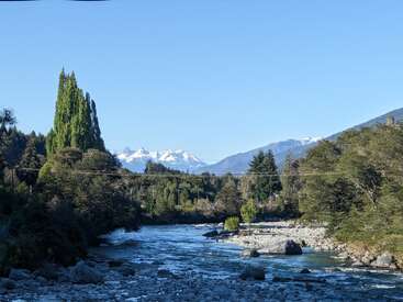 Une rivière sereine coule dans une vallée verdoyante, entourée de grands arbres, avec au loin des montagnes enneigées sous un ciel bleu clair. Scène de nature paisible.