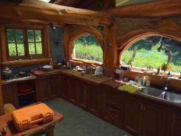 Essa cozinha rústica aconchegante apresenta vigas de madeira, armários de madeira, luz natural de grandes janelas e vários itens essenciais de cozinha espalhados nas bancadas, criando uma atmosfera calorosa e convidativa.