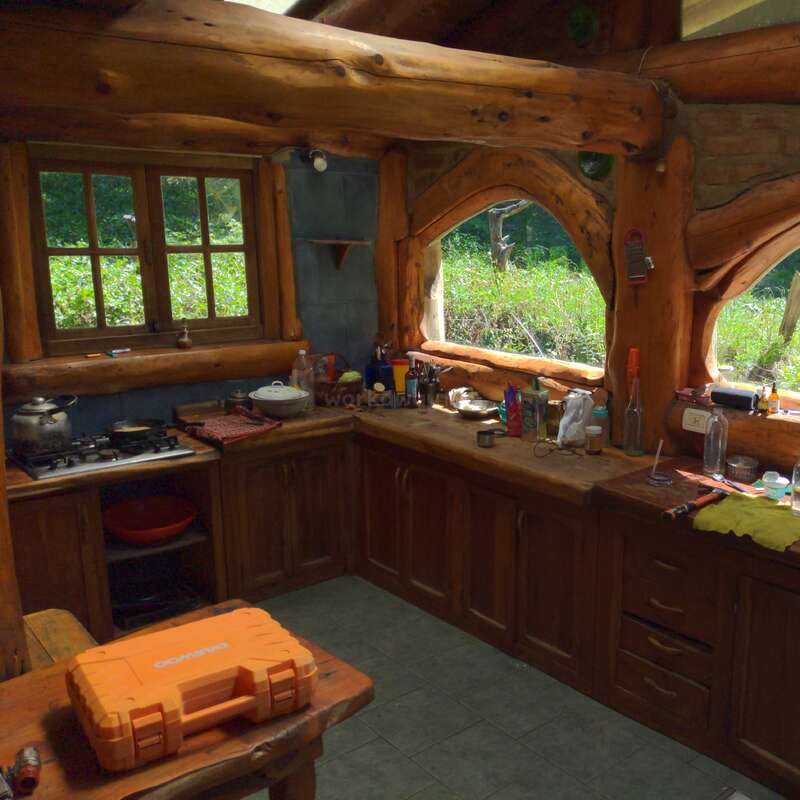 This cozy rustic kitchen features log cabin beams, wooden cabinets, natural light from large windows, and various kitchen essentials scattered on countertops, creating a warm, inviting atmosphere.