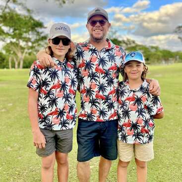 A imagem mostra um homem e dois meninos juntos em um campo de golfe, todos vestindo camisetas iguais com palmeiras e flamingos.