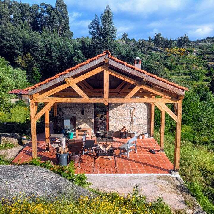 Das Bild zeigt einen ruhigen Terrassenbereich mit einer hölzernen Pergola, umgeben von üppigem Grün und einem blauen Himmel mit weißen Wolken.