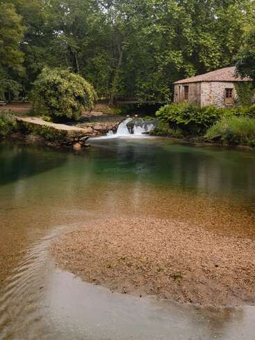 Ein ruhiger Fluss fließt sanft an einem alten Steinhaus vorbei, mit einem kleinen Wasserfall, üppigem Grün, einem sandigen Ufer und einer friedlichen Waldkulisse.
