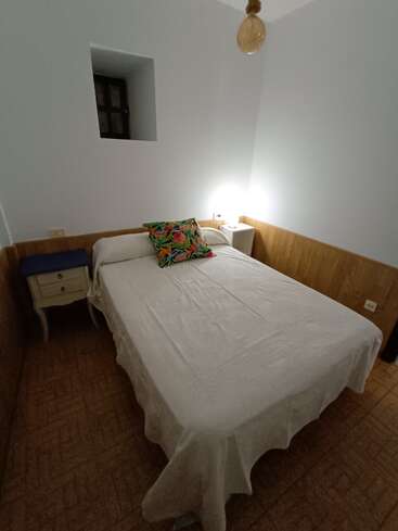 Ein kleines, gemütliches Schlafzimmer mit einem Doppelbett, weißer Bettwäsche, bunten Kissen, zwei Nachttischen, einer Lampe, einem kleinen Fenster, hellen Wänden und einer gewebten Deckenleuchte.