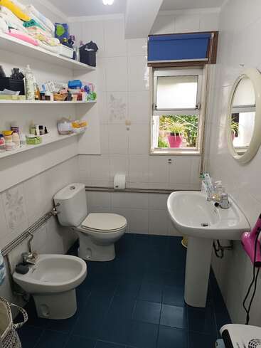 Es ist ein aufgeräumtes Badezimmer mit blauem Fliesenboden, weißen Armaturen, offenen Regalen voller Toilettenartikel und Handtücher, einem Fenster und einem runden Spiegel über dem Waschbecken.