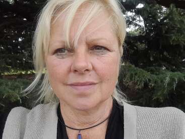 A middle-aged woman with short blonde hair takes a selfie outdoors. She wears a gray sweater, black top, necklace, and stands in front of green trees.