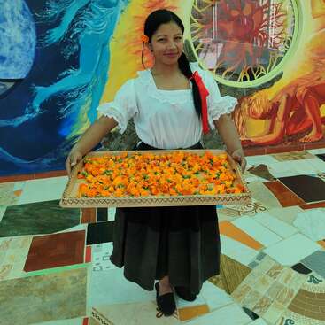 Eine Frau in traditioneller Kleidung hält ein Tablett mit leuchtend orangefarbenen Blumen und steht vor einem farbenfrohen Wandgemälde mit blauen, gelben und roten Farbtönen und Figuren.