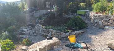 Uma pessoa cuida de um jardim com terraços de pedra. Há plantas verdes, um balde amarelo, uma banheira verde e ferramentas de jardinagem e pedras espalhadas.