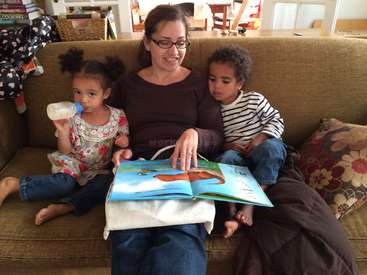 A imagem mostra uma mulher lendo um livro para duas crianças em um sofá, com a mulher segurando uma mamadeira e as crianças sentadas em cada lado dela.