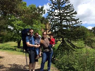 A imagem mostra uma família de quatro pessoas posando para uma foto em um parque, com o pai carregando uma criança nas costas e a mãe segurando outra criança.