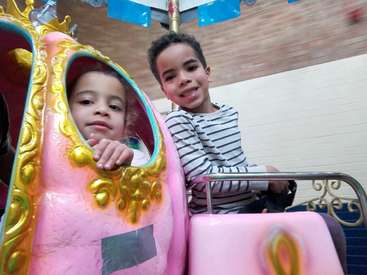A imagem mostra duas crianças andando em um carrossel rosa e dourado, com o menino à direita e a menina à esquerda, ambos sorrindo e olhando para a câmera.