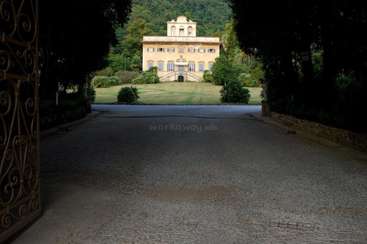 The image depicts a large, yellow house with a driveway and a stone wall, set against a backdrop of trees and a hill.