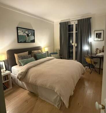 A cozy bedroom with a neatly made bed, decorative pillows, bedside lamps, a desk with a chair, curtained window, wooden floor, and artwork on the walls.