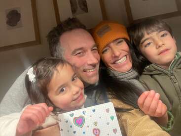 A joyful family selfie with two adults and two children, all smiling warmly. The girl in front holds a drawing decorated with colorful hearts. Cozy atmosphere.