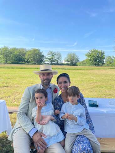 A family of four poses outside on a sunny day. The father wears a light suit and hat, the mother a blue dress, children look curious.