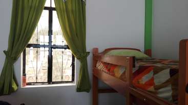 La imagen muestra una habitación con cama, escritorio y silla, y una ventana con vistas a árboles y una masa de agua al fondo. La habitación está bien iluminada y tiene una combinación de colores neutros.