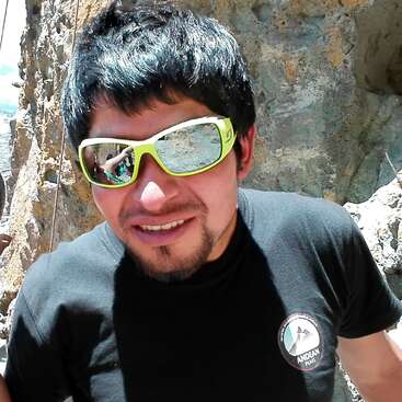 La imagen muestra a un hombre de pelo oscuro y camiseta negra, de pie frente a un acantilado rocoso, con gafas de sol y bigote y perilla.