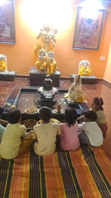 Cinco niños sentados en una estera a rayas en un templo, frente a deidades hindúes y un lingam de Shiva, preparando u ofreciendo prasad en un entorno ritual.