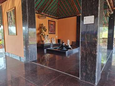 Esta imagen muestra un sereno templo interior con pilares de mármol, estatuas de deidades hindúes, arte religioso enmarcado, un Shiva Lingam, suelos pulidos y un tejado de tejas verdes. Ambiente apacible.