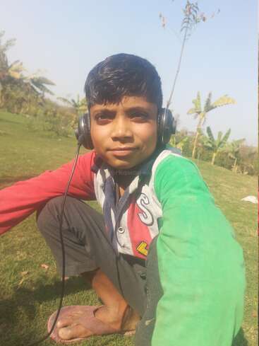 Un niño está sentado al aire libre sobre hierba verde, con auriculares y una chaqueta de colores. Hace sol, está rodeado de árboles y se ve un cielo azul despejado.