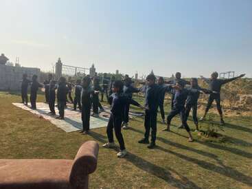 Un grupo de niños, vestidos con uniformes oscuros, están de pie al aire libre sobre la hierba, realizando ejercicios o yoga con los brazos extendidos bajo el brillante cielo soleado.
