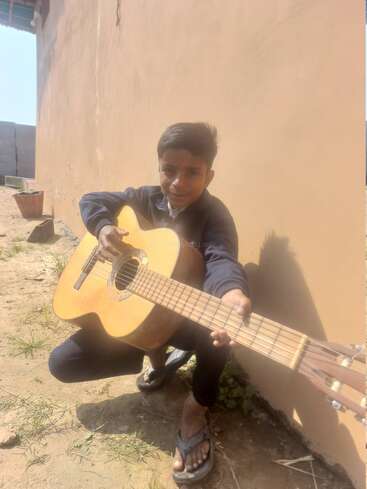 Un joven está en cuclillas junto a una pared, sonriendo mientras sostiene y toca una guitarra acústica. La luz del sol ilumina la escena, resaltando su atuendo informal y sus sandalias.