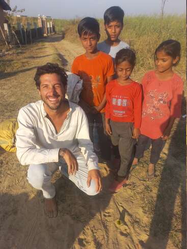 Un hombre está en cuclillas y sonríe alegremente en un soleado camino de tierra, rodeado de cuatro niños. El grupo parece feliz al aire libre, con un paisaje rural de fondo.