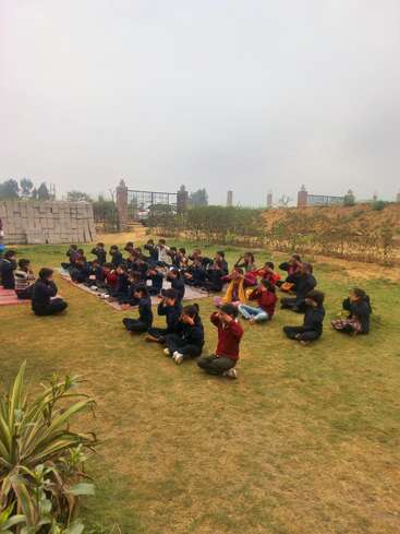 Un grupo de niños sentados al aire libre en colchonetas, realizando una actividad o meditación en grupo, con vegetación, ladrillos y una verja de fondo.