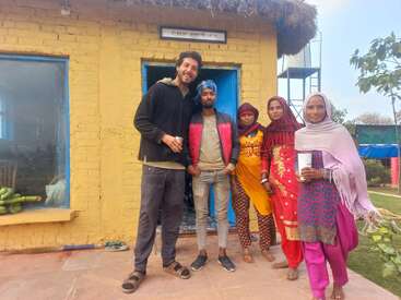 Cinco personas permanecen juntas delante de una casa amarilla con puertas y ventanas azules. Parecen felices, posando para una foto de grupo con ropa informal.