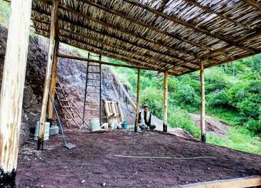 A man sits under a simple bamboo and wood shelter under construction in a green, hilly area. Tools, buckets, and ladders are nearby, showing ongoing work.