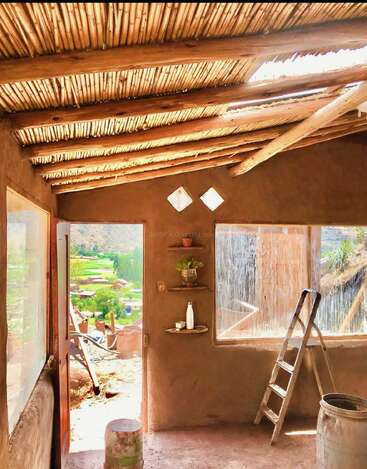 Sunlit rustic room with bamboo roof, open door, and large windows. Simple shelves hold plants and a bottle. Ladder and barrel add charm, overlooking scenic greenery.