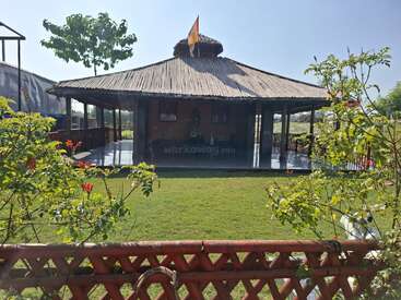 Ein strohgedeckter offener Pavillon steht in einem üppigen grünen Garten, umgeben von Pflanzen und einem Holzzaun. Auf dem Gebäude ist eine Flagge unter blauem Himmel zu sehen.