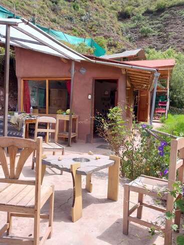 Une terrasse rustique, avec des chaises en bois et une table en pierre, jouxte une maison de style adobe. Une végétation luxuriante, des collines et des fleurs aux couleurs vives viennent compléter ce tableau paisible.