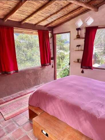 Cette chambre chaleureuse est dotée d'un plafond rustique en bois, de grandes fenêtres ornées de rideaux rouges, d'un couvre-lit rose, de murs aux tons ocres, d'un sol carrelé et offre une vue imprenable sur la verdure.