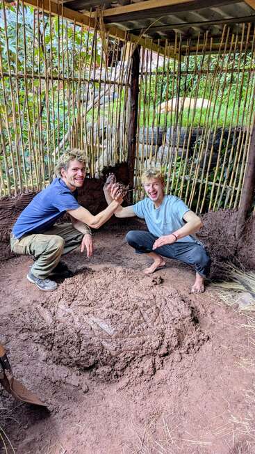 Deux jeunes hommes souriants, les mains couvertes de boue, sont agenouillés à l'intérieur d'un abri en bambou. Ils se réjouissent d'avoir travaillé ensemble à un projet en terre, au milieu d'un cadre naturel verdoyant.
