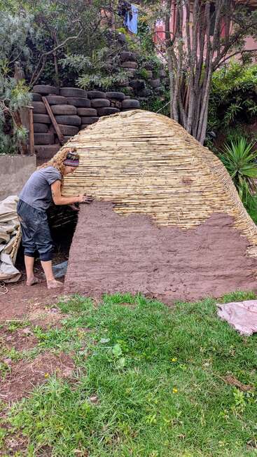 Une personne applique de la boue sur une structure en bambou, probablement pour construire une maison en terre. À l'arrière-plan, on aperçoit de la végétation, des pneus empilés et une maison partiellement visible.