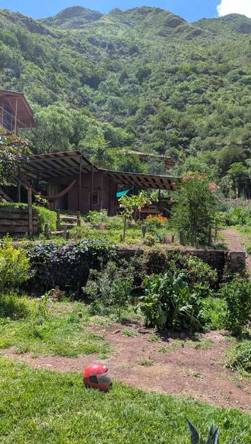 Une cabane rustique équipée de hamacs se niche dans une vallée verdoyante, entourée de montagnes et de jardins. Un casque rouge repose sur la pelouse d'un vert éclatant.