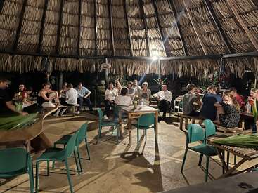 Eine Gruppe von Menschen speist und unterhält sich abends unter einem großen, strohgedeckten Pavillon, umgeben von Holztischen, grünen Stühlen und weicher, warmer Beleuchtung.