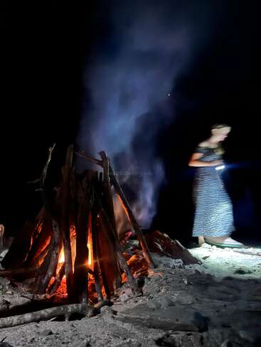 Nachts brennt ein Lagerfeuer hell auf dem sandigen Boden und lässt Rauch in die Höhe steigen. Eine Person in einem langen Kleid geht vorbei und hält eine Taschenlampe in der Hand.