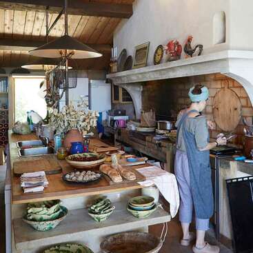 L'image représente une femme en train de cuisiner dans une cuisine rustique, entourée de comptoirs en bois, d'un four en briques et d'un assortiment de plats et d'ustensiles.