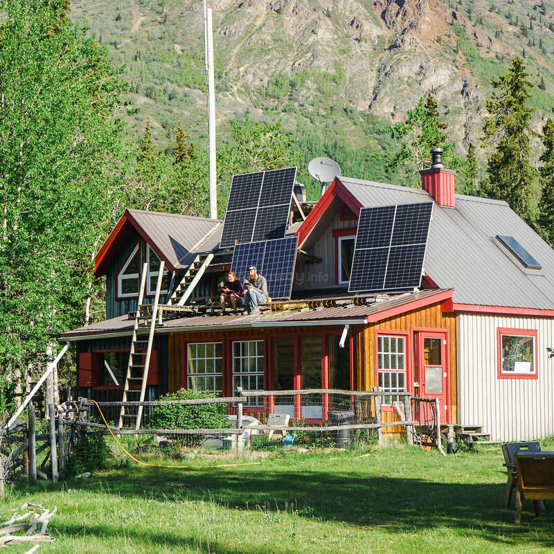 Two people sit on a house roof surrounded by trees and mountains. Solar panels are installed on the roof, and a ladder leans against the house.